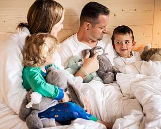 Eine Familie entspannt im Bett mit mehreren Kuscheltieren.