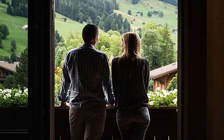 Ein Paar steht am Balkon und blickt auf eine grüne Hügellandschaft.