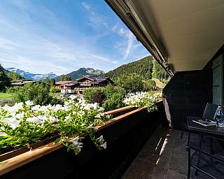 Balkon mit weißen Blumen, Tisch und Stühlen sowie Blick auf Berge und Häuser im Tal; the content_suitable ist true.